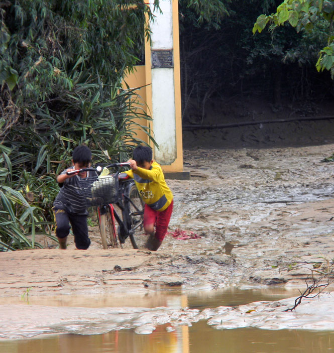 Hai em nhỏ ở xã Tiến Hoá đang nỗ lực dắt xe đạp qua đoạn đường ngập bùn đất sau lũ.